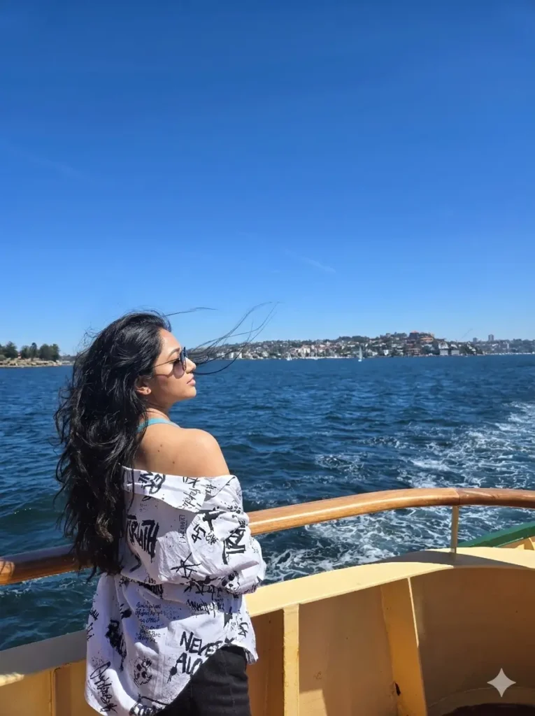Person with long dark hair wearing sunglasses, standing on a boat deck and gazing at the water under a clear blue sky.