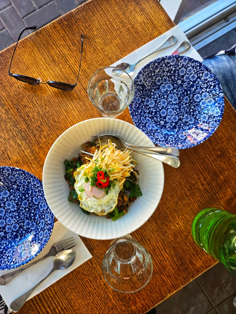 Top-down view of a wooden table with a bowl of food topped with a fried egg, bean sprouts, and greens, surrounded by blue patterned plates, cutlery, glasses, and sunglasses in natural sunlight.