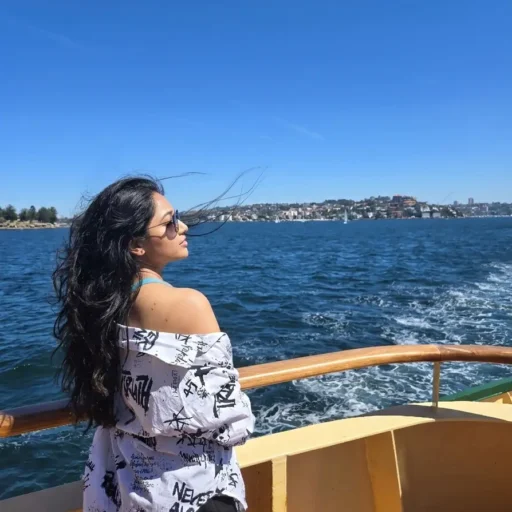 Person with long dark hair wearing sunglasses, standing on a boat deck and gazing at the water under a clear blue sky.