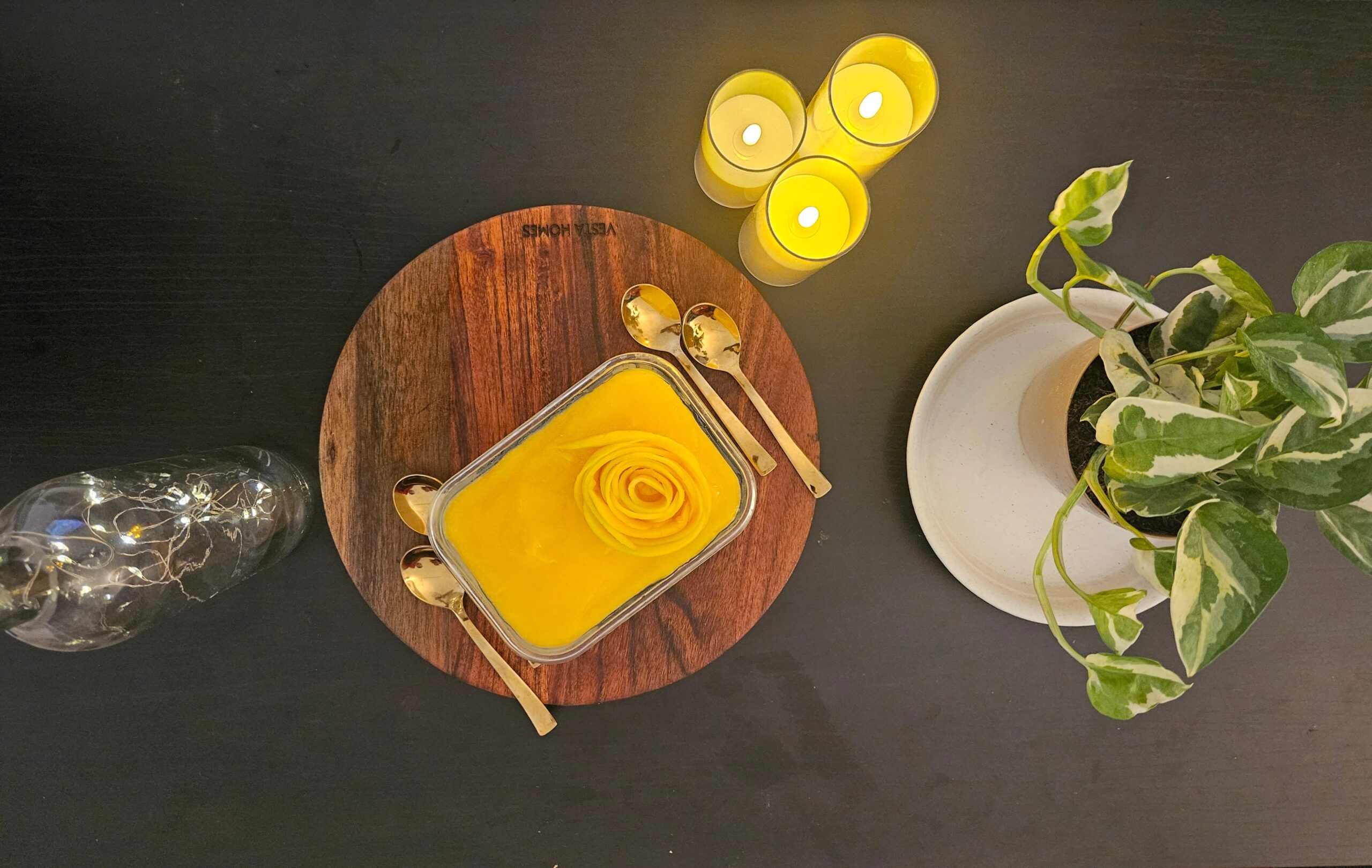 Top view of a healthy no-bake mango cheesecake with a mango rose garnish served on a wooden board beside candles and indoor plants