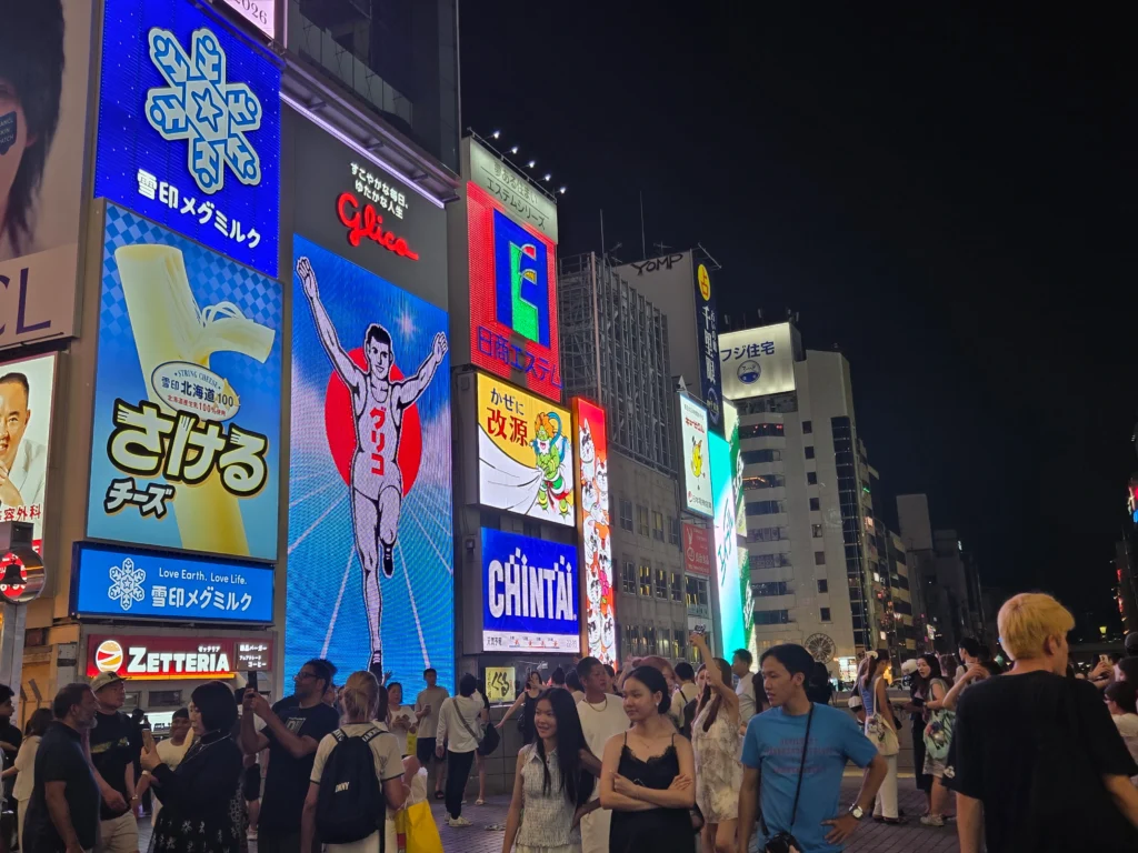 Crowds gathered in Osaka’s Dotonbori in July at night beneath brightly lit billboards, including the famous Glico Running Man sign, with colorful neon lights illuminating the street.
