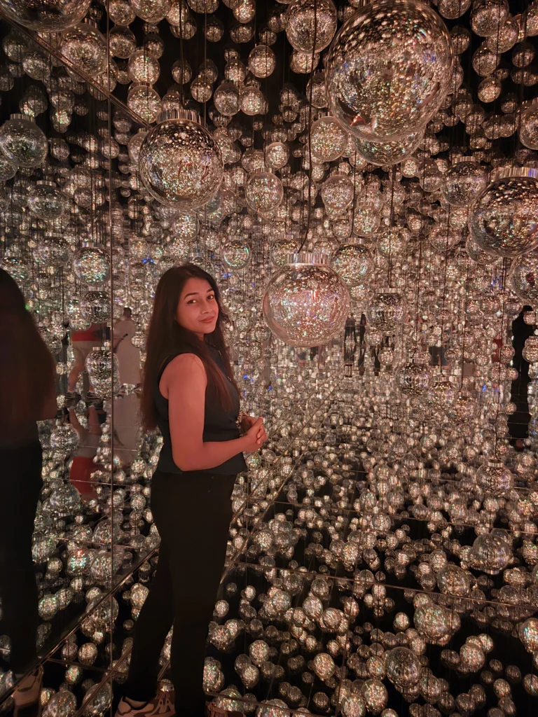 Nikita standing inside a mirrored infinity room installation filled with reflective glass orbs, surrounded by shimmering light and endless reflections