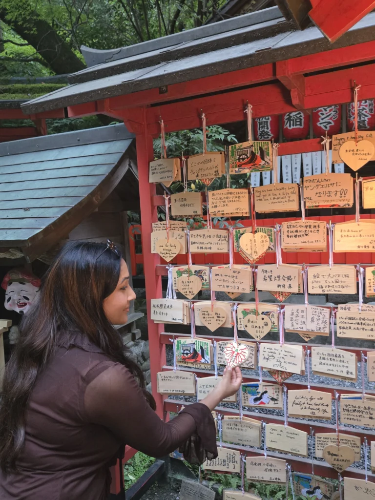 Nikita reading wooden ema wish plaques hanging at a Japanese shrine, surrounded by red torii-style architecture and hanging lanterns