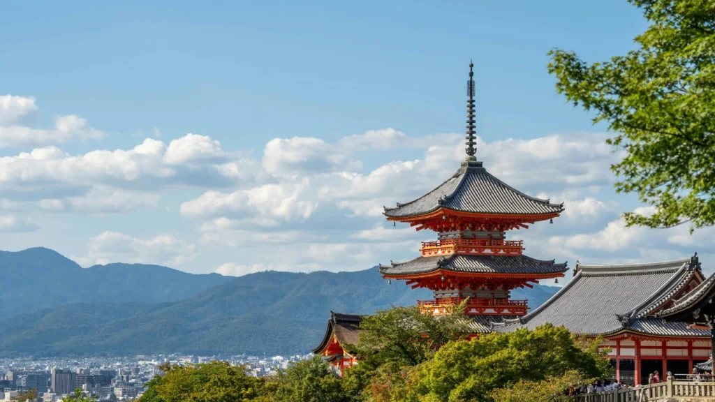 A view of the iconic three-story red and orange pagoda at Kiyomizu-dera temple in Kyoto, Japan, set against green trees, mountains, and a clear blue sky with fluffy white clouds.