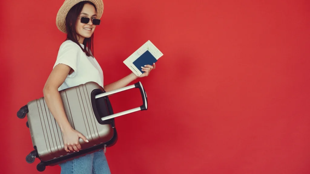 A smiling young woman wearing a straw hat, sunglasses, and a white t-shirt, carrying a grey rolling suitcase while holding a map and passport against a bright red background.